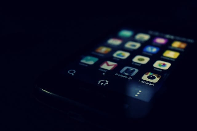 В темном фоне виден смартфон с включенным экраном. На экране отображены различные иконки приложений, в том числе иконка Instagram. Внизу экрана есть кнопки навигации.