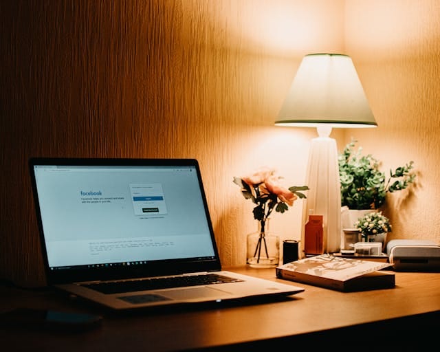 Một chiếc máy tính xách tay được mở trên bàn, màn hình hiển thị trang đăng nhập của Facebook. Gần máy tính có một cái đèn nệm sáng lên, bên cạnh đó là một chai hoa và một số đồ dùng khác trên bàn.
