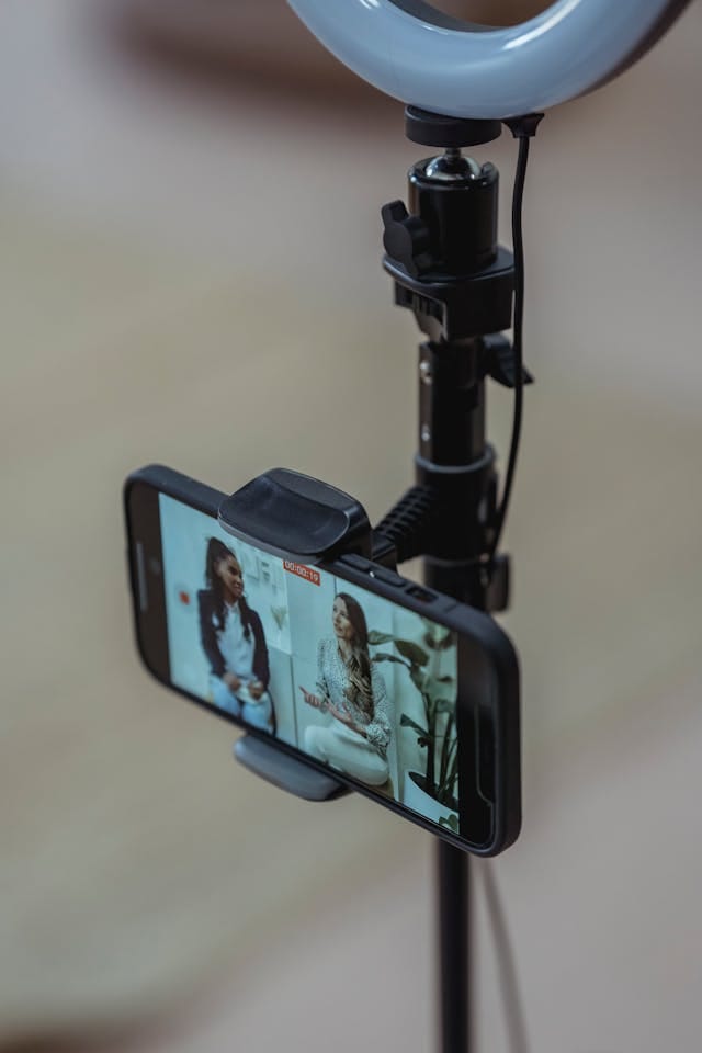 Sebuah ponsel terpasang kokoh di dudukannya, disertai dengan ring light yang bersinar terang di atasnya. Pada layar ponsel, terlihat sebuah video yang menampilkan dua orang sedang berbincang dengan ekspresi penuh antusiasme.
