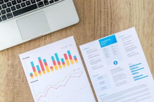Sebuah laptop terletak di atas meja, bersama - sama dengan beberapa lembar kertas yang menampilkan grafik bisnis dan sebuah resume. Di era digital saat ini, data dan analisis seperti yang terlihat di kertas - kertas ini dapat membantu para pengguna Facebook untuk memahami dan meningkatkan performa konten mereka di platform sosial media tersebut.