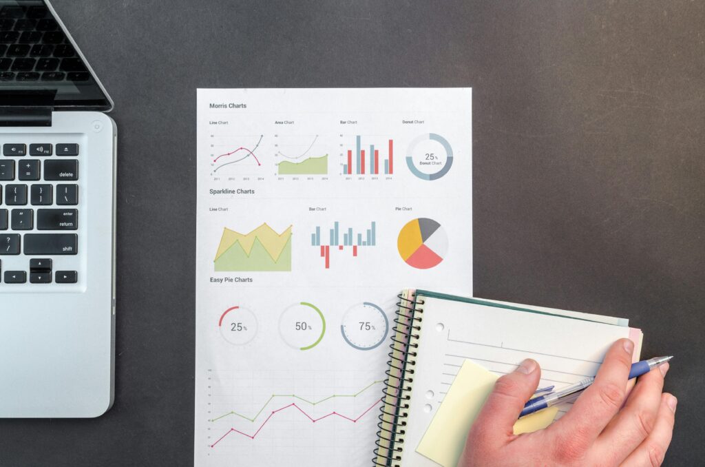 Sebuah meja dengan laptop, selembar kertas yang menampilkan berbagai jenis diagram seperti grafik garis, diagram batang, dan diagram lingkaran. Di sampingnya ada buku catatan dan pensil. Ini bisa menjadi sumber inspirasi untuk membuat konten educatif tentang data dan analisis di TikTok.