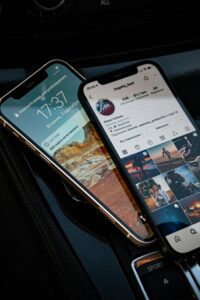 Two smartphones are placed on a car's surface. One displays a lock - screen with a scenic wallpaper and time, while the other shows a social media profile. Such devices are often used for apps like ShareChat to connect with others and share content.