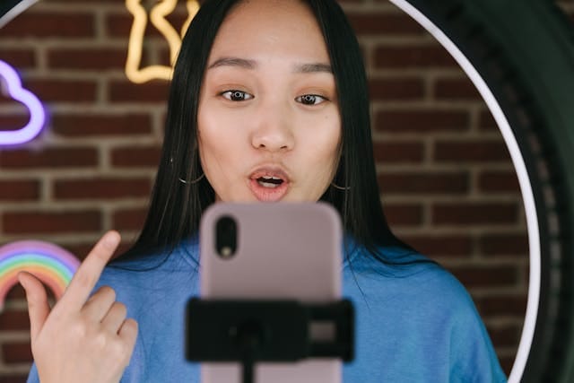 Una mujer con cabello largo y un top azul está delante de una iluminación de anillo, apuntando un teléfono móvil hacia sí misma. Detrás de ella, hay una pared de ladrillos con luces de neón en forma de arcoíris y otras formas. Hace un gesto con el dedo.