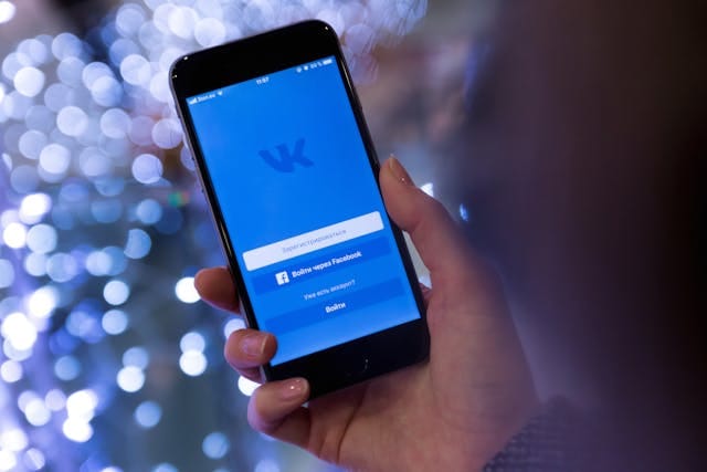 Eine Hand hält ein Mobiltelefon mit einem blauen Display, auf dem das Logo von VK sichtbar ist. Auf dem Bildschirm werden Optionen wie "Login mit Facebook" und "Weitere Optionen" angezeigt. Im Hintergrund sind unscharfe, beleuchtete Kugeln zu sehen, die einen weihnachtlichen oder festlichen Eindruck vermitteln.