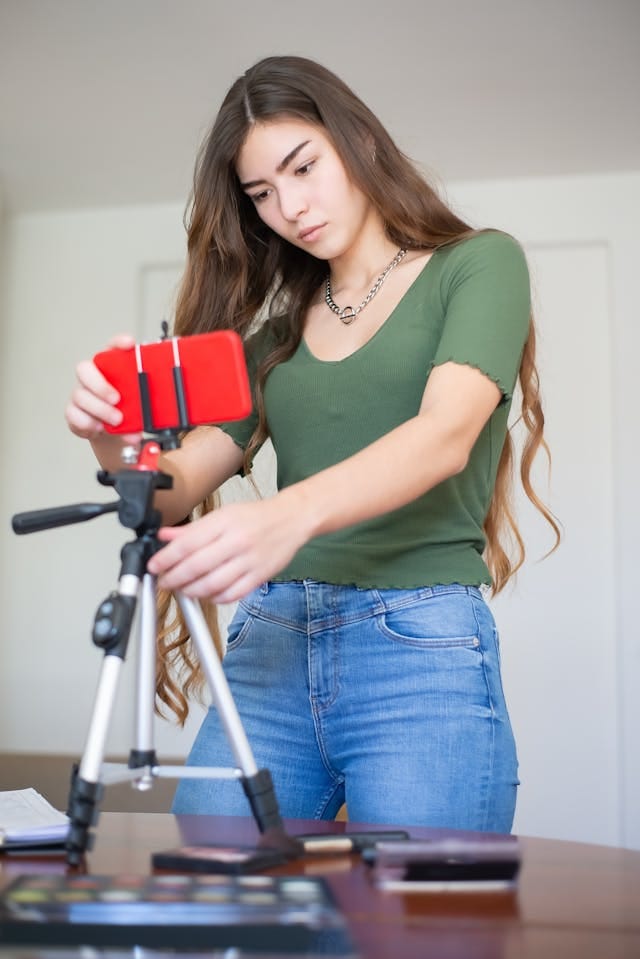Eine junge Frau mit langem, braunem Haar trägt ein grünes, kurzärmliges Shirt und blaue Jeans. Sie befestigt ein rotes Mobiltelefon in einem Halter auf einem dreibeinigen Stativ. Auf dem Tisch vor ihr befinden sich verschiedene Gegenstände, darunter Makeup - Produkten, was darauf hindeutet, dass sie möglicherweise eine Videoblog - Session vorbereitet.