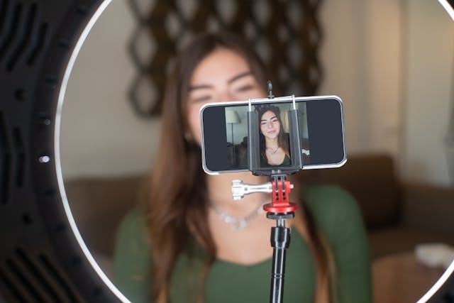 Eine junge Frau mit langem, braunem Haar sitzt hinter einem Mobiltelefon, das auf einem Stativ befestigt ist. Das Telefon zeigt ein Live - Bild von ihr. Ein Ringlicht umgibt das Telefon und beleuchtet sie. Im Hintergrund ist eine Couch und eine Wanddekoration zu sehen.