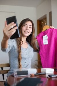 Eine junge Frau mit langem, braunem Haar hält ein schwarzes Mobiltelefon in ihrer linken Hand, um sich ein Selfie zu machen. Mit ihrer rechten Hand hält sie ein pinkes, nagellanges Oberteil an einem Holzhaken. Auf dem Tisch vor ihr befinden sich verschiedene kosmetische Artikel wie Puder und Pinseln.