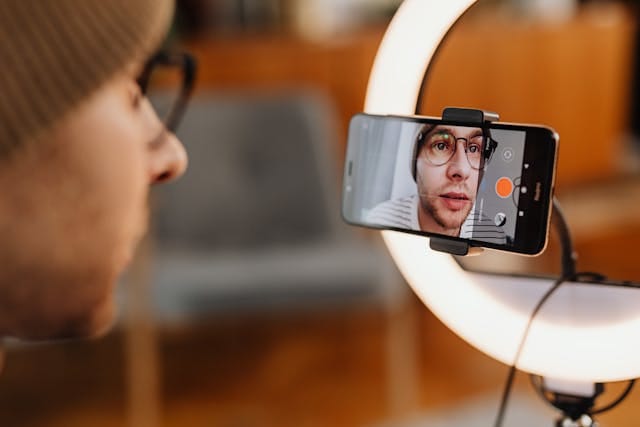 Un individu avec un bonnet et des lunettes se filme avec un téléphone portable monté sur un support. Un anneau - lumière éclaire la scène. L'écran du téléphone affiche l'image de la personne en train de filmer.