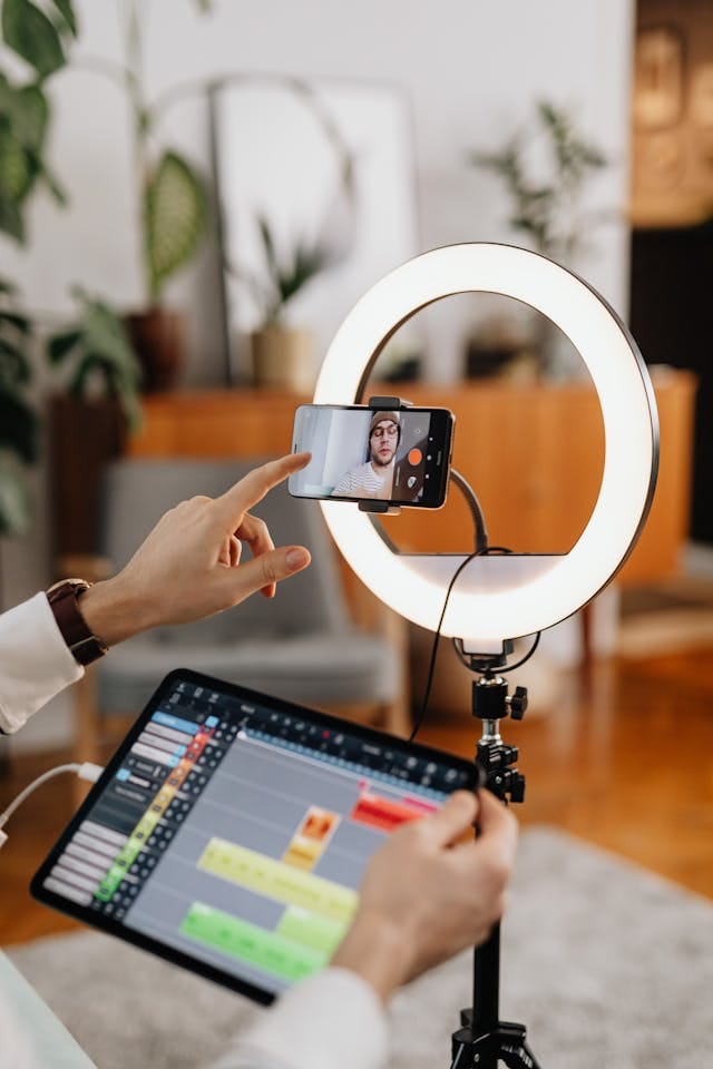 Des mains manipulent un téléphone portable placé sur un support, avec un anneau - lumière éclairant la scène. L'écran du téléphone affiche une image de quelqu'un. En même temps, une tablette est tenue, affichant une interface de montage vidéo avec des bandes de médias de différentes couleurs. L'arrière - plan est décoré avec des plantes et des meubles.