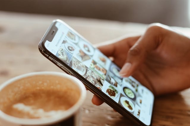 手が、画面に料理の写真が並んだスマートフォンを持っています。スマートフォンの横には、コーヒーが入ったコップが置かれています。