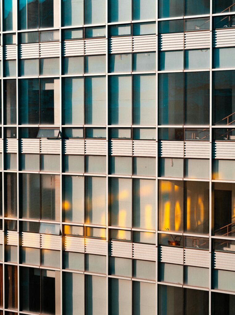 Ein modernes Hochhaus mit einer Fassade aus Glas - und Metall - Elementen. Die Glasscheiben reflektieren das Umfeld, darunter gelbe Lichter und Teile anderer Gebäude, während die horizontalen Metallbalken die Architektur strukturieren.