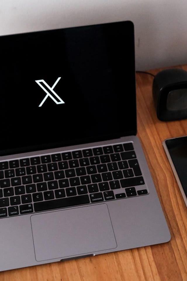 مكتب حديث بتصميم رمادي أنيق، تتوسطه شاشة سوداء بارزة تحمل **رمز "X" باللون الأبيض**، مما يضفي لمسة تقنية عصرية. إلى الجانب الأيمن من الشاشة، **يوجد جهاز إلكتروني صغير**، يكمل المشهد بإحساس من **التطور والابتكار**، بينما يعكس الترتيب الدقيق للمكتب **بيئة عمل احترافية ومتناغمة**.