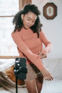 A woman in a peach - colored knitted sweater is standing in a cozy room, adjusting her sleeve in front of a camera. Platforms like VKontakte could be used to share similar content of fashion and lifestyle moments.