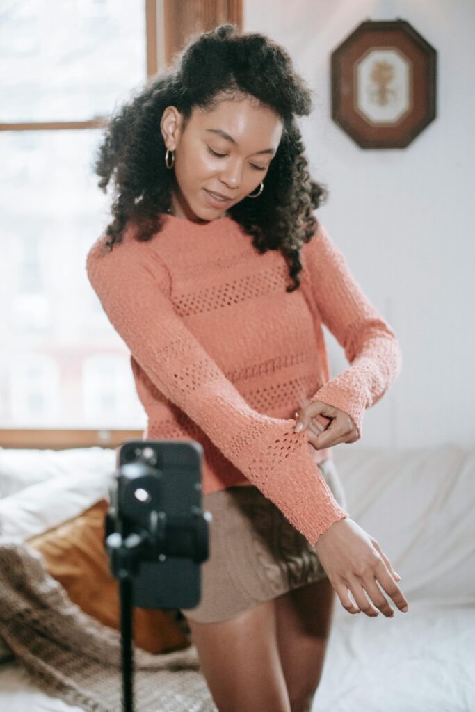 A woman in a peach - colored knitted sweater is standing in a cozy room, adjusting her sleeve in front of a camera. Platforms like VKontakte could be used to share similar content of fashion and lifestyle moments.