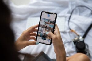 2 つの手が、画面に写真が表示されたスマートフォンを持っています。手の爪にはネイルアートが施されており、背景には白い布、カメラやキーボードのようなものが见えます。