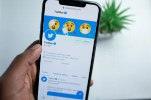 A hand - held smartphone displays the Twitter app interface, featuring the Twitter logo, a profile picture, and emojis related to health and safety on the profile page.