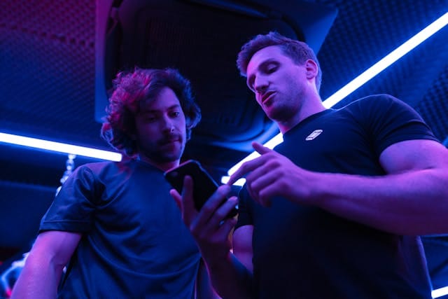 Dois homens em uma iluminação com tons de azul e roxo estão olhando para um celular, com um deles apontando para a tela.