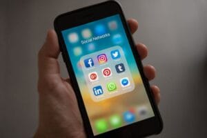 يد تحمل هاتفًا محمولًا يظهر على شاشته كلمتا "Social Networks" وآيقونات لبرامج التواصل الاجتماعي مثل Facebook، Instagram، Twitter، TikTok، Pinterest، LinkedIn و WhatsApp.
