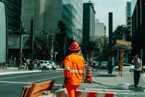 A worker in an orange safety suit and red hard hat stands on a busy urban street in Brazil. Surrounding are modern buildings, pedestrians, and vehicles. In the context of 2025 Facebook Influencer Marketing Trends in Brazil, this scene could represent the blend of everyday life and potential marketing opportunities.