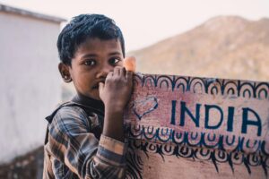 A young boy in a plaid shirt is peeking out from behind a board with "INDIA" written on it, with mountains in the background. 2025 Social Media Marketing Outlook in India - This image captures the essence of India and can inspire creative social media marketing strategies in the country in 2025, highlighting its unique people and landscapes.