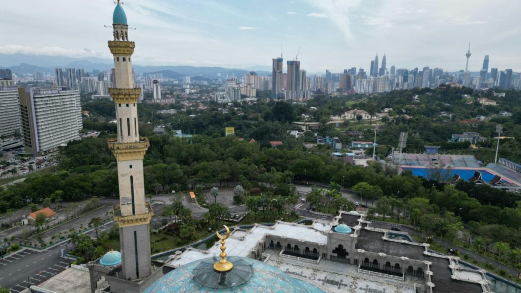 Sebuah masjid dengan tiang minaret yang menjulang tinggi dan kubah berwarna biru, dikelilingi oleh pepohonan hijau. Di latar belakangnya adalah panorama kota dengan gedung - gedung menakjubkan. Di tengah keindahan ini, muncul pertanyaan: Apa Kabar Dubsmash Aplikasi Lipsync yang Dulu Viral Sebelum TikTok Lahi