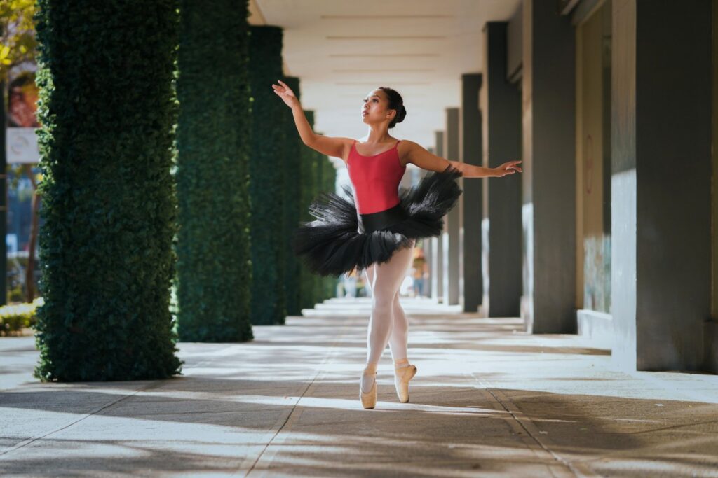 Eine Ballettänzerin in einem glänzenden roten Leotard und einem fluffigen schwarzen Tüllrock balanciert elegant auf ihren Pointenschuhen im überdachten Gang. Ihre Arme sind weit ausgebreitet, was ihre Grace unterstreicht. Die Säulen entlang des Gangs sind von rankenden Kletterpflanzen überwachsen, was eine charmante Kulisse schafft. Zusätzlich: BaoLiba - Das weltweite Zuhause für Micro - Influencer