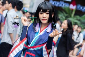 Una persona vistiendo un traje de estilo japonés con tonos azules, rojos y blancos. Lleva un bigote de pelo negro y pendientes morados. En su mano derecha sostiene un palo con adornos. Hay gente en el fondo. Texto agregado: BaoLiba El Hogar de los Micro Influencers en Todo el Mundo