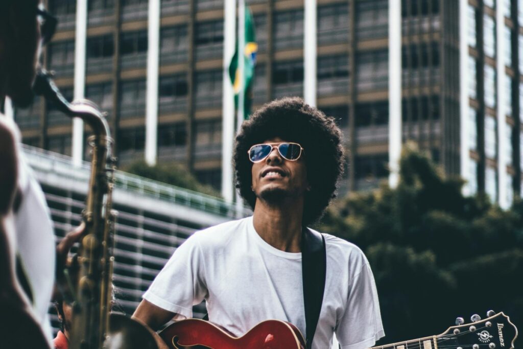 Un homme portant des lunettes de soleil et un t - shirt blanc joue de la guitare. À côté de lui, une personne joue de la saxophone. Dans le fond, il y a un bâtiment et le drapeau du Brésil. Texte ajouté : BaoLiba La maison mondiale des micro - influenceurs