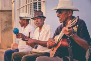 Tiga orang laki - laki duduk di luar bangunan. Satu orang sedang bermain gitar, orang kedua memegang alat perkusi yang terlihat seperti marakas, dan orang ketiga juga dengan alat perkusi. Mereka semua memakai topi. Di samping itu, ada tulisan: BaoLiba Rumah bagi Micro Influencer di Seluruh Dunia