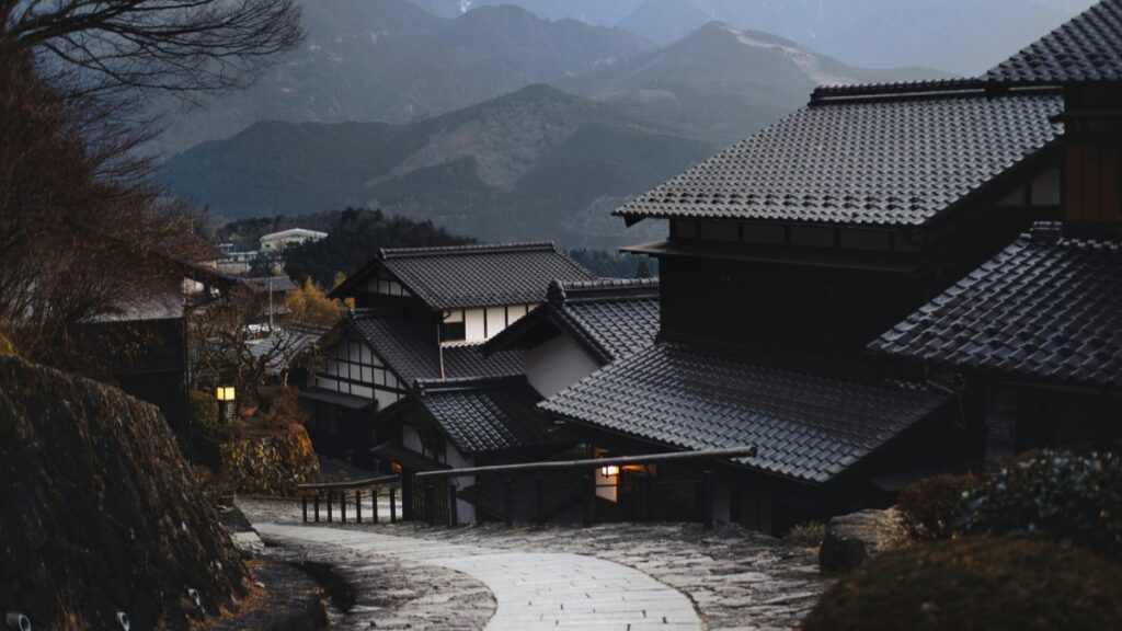 この画像は、日本の伝統的な集落を描いています。瓦屋根の家屋が連なり、背後には山々が広がっています。小道には灯りがともり、落ち着いた雰囲気が漂っています。このような美しい風景を楽しむだけでなく、デジタル時代においては、Bigo Liveでお金を稼ぐには？ということも関心の対象となっています。
