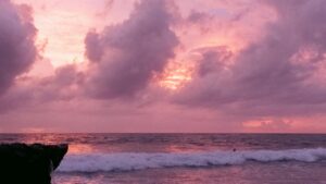 Suasana pantai saat matahari terbenam, langit dipenuhi awan yang berwarna merah muda sampai ungu, dan ombak-ombak melambung ke pantai. Di tengah keindahan alam ini, muncul pertanyaan: Cara Menghasilkan Uang dari Grup Facebook