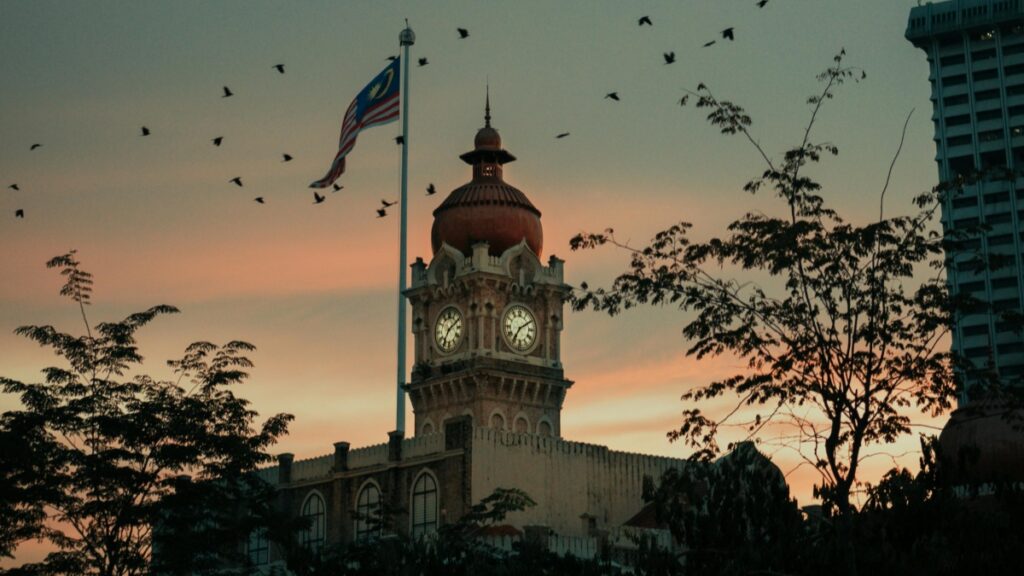 Gambar ini menunjukkan sebuah gedung dengan menara jam yang mempesona, dengan bendera Malaysia yang berkibar dengan semangat. Di latar belakang, langit berwarna merah keangsa saat senja, dan sejumlah burung terbang mengelilinginya. Selain menikmati pemandangan indah seperti ini, bagi mereka yang tertarik dengan dunia digital, belajar Cara Menghasilkan Uang di Bigo Live dapat menjadi pilihan untuk memperoleh pendapatan tambahan.