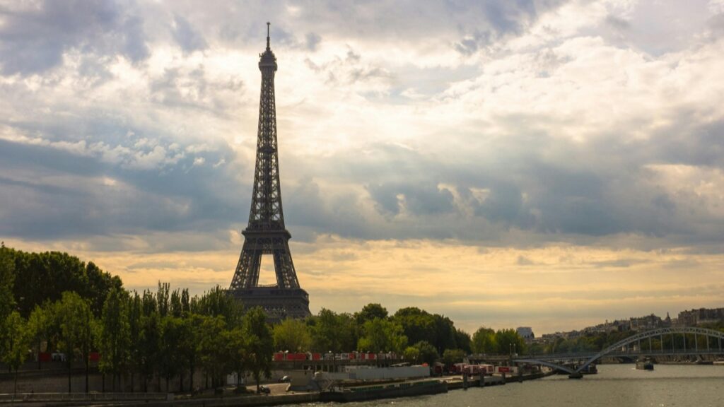 Le célèbre Tour Eiffel se dresse majestueusement au milieu de la végétation, avec le fleuve en avant - plan et un pont dans la distance. Le ciel est couvert de nuages. Face à cette vue emblématique de Paris, une question vient à l'esprit : Comment Gagner de l’Argent avec un Groupe Facebook ?