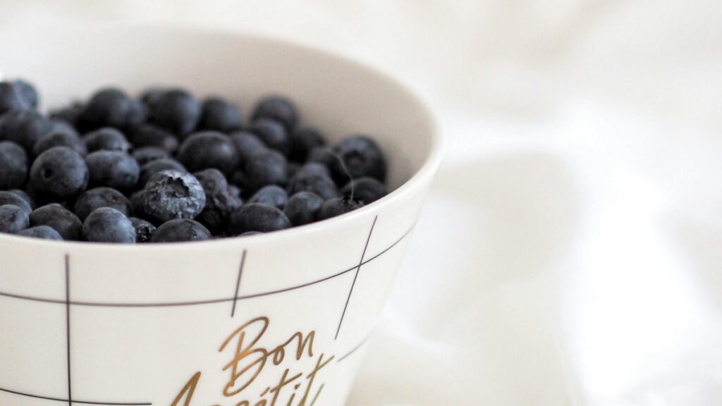Un bol blanc avec un motif de lignes et les mots "Bon Appétit" écrits en lettres ornementales. Il est rempli de myrtilles bleues sombres. Bien que les myrtilles soient un délicieux snack santé, on peut également voir une analogie avec "Comment Gagner de l'Argent sur Signal", en termes de la nécessité de choisir avec soin, tout comme on choisit des myrtilles fraîches, pour avoir des résultats fructueux sur le plan financier.