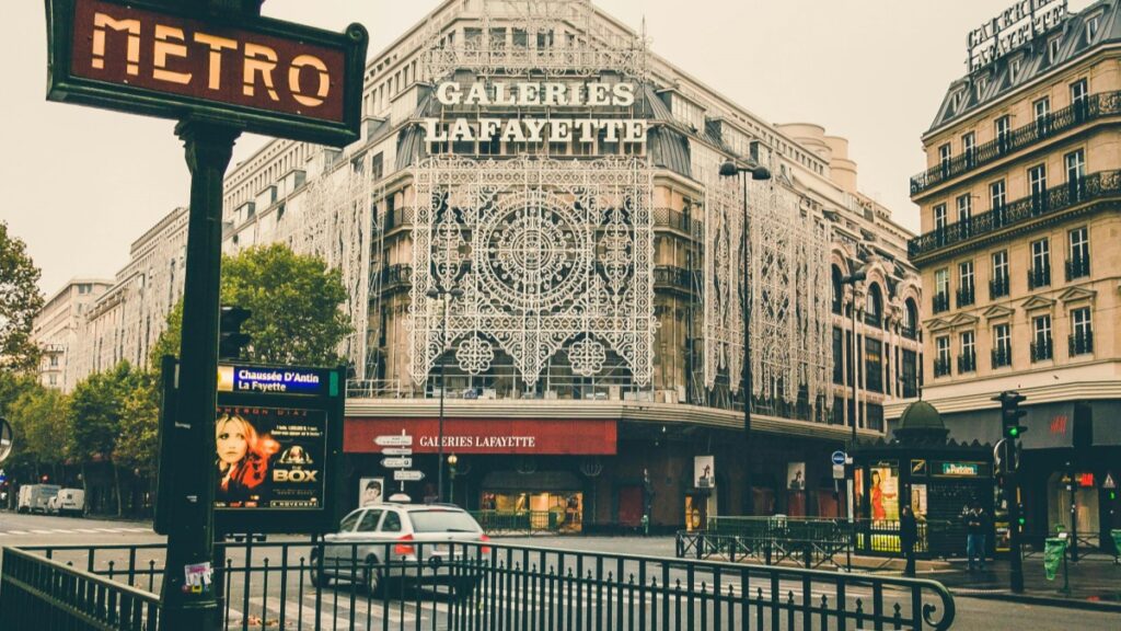 Dans cette image, on voit le célèbre magasin Galeries Lafayette à Paris, avec un panneau "METRO" en avant-plan. La façade du magasin est ornée d'un élégant motif. Autour, il y a des voitures, des panneaux publicitaires et d'autres immeubles. Alors que vous explorez les lieux emblématiques de Paris, vous pouvez également être intéressé par Comment gagner de l'argent sur Bigo Live, une opportunité pour développer votre revenu en ligne.