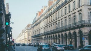 Una calle en una ciudad con edificios antiguos y elegantes en ambos lados. Hay varios coches en la calle y gente caminando. Un semáforo muestra la luz verde. Mientras se ve esta escena urbana, surge la pregunta: ¿Cómo Ganar Dinero con Grupos de Facebook?