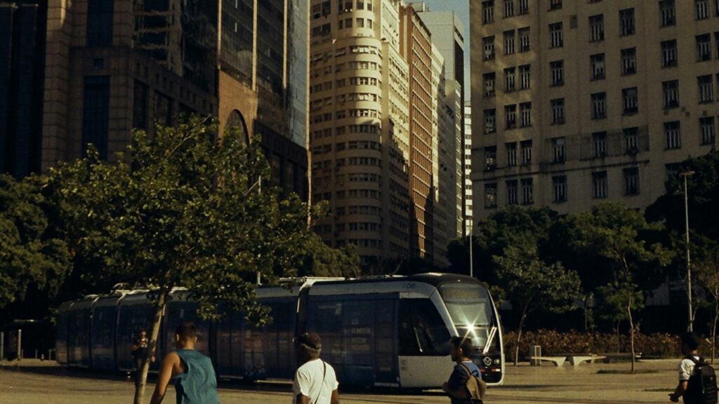 Na cena, um trem de rua em movimento com edifícios altos ao fundo. Pessoas caminham na área, com algumas árvores na paisagem. Diante desse cenário urbano, surge a pergunta: Como Ganhar Dinheiro com Grupos do Facebook no Brasil.