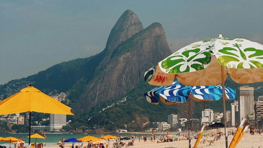 Uma vista paradisíaca da praia, com um monte emblemático no fundo. As praias estão cheias de guarda - sol coloridos, incluindo amarelos, azuis e brancos com padrões verdes. Há muitas pessoas curtindo o sol e o mar. É um cenário vibrante e convidativo. E, Como Ganhar Dinheiro no Instagram no Brasil (de verdade, não no papo furado)