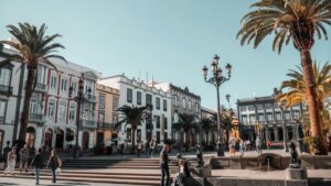 Una plaza animada con edificios coloniales de estilo antiguo, rodeados de palmeras altas y con una atmósfera soleada. Gente camina y se detiene, disfrutando del día. Al igual que esta plaza es un lugar de encuentro y actividad, "Cómo ganar dinero en Tinder en España (sin estafas, sin postureo y sin vender humo)" ofrece la posibilidad de descubrir oportunidades financieras legítimas en el mundo digital.