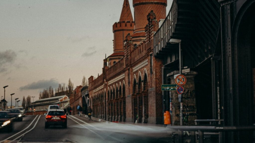 In diesem Bild ist eine Straße zu sehen, an der ein imposanter, rot - backsteiniger Bau mit turmförmigen Zügen steht. Autos fahren auf der Straße, und an der Straßenseite befinden sich Verkehrsschilder. Die Stimmung ist von einer düsteren, trüben Atmosphäre geprägt. Und in der Tech - Szene bringt Firework das erste Live - Shopping - GPT der Welt, was sicherlich die E - Commerce - Welt verändern wird.