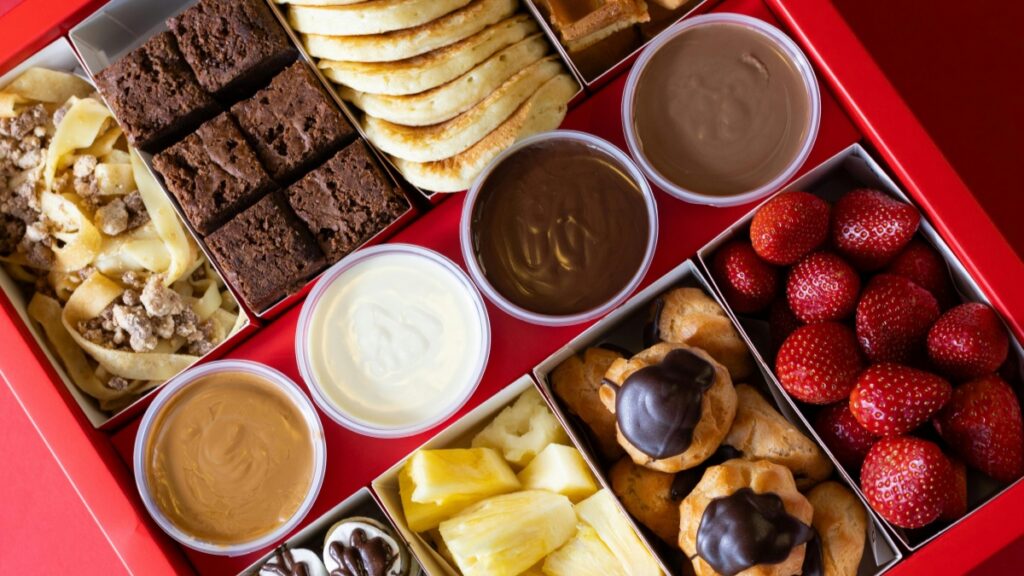 Kotak makanan penutup merah yang berisi berbagai makanan manis seperti brownies, krem, susu stroberi, dan waffle. Di sampingnya ada gelas coklat dan krim yang mengalir. Menghasilkan Uang dengan Funimate, Panduan Praktis untuk Kreator Indonesia.