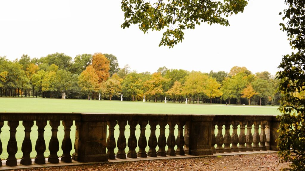 Eine wunderschöne Ansicht eines Parks mit einem großen Rasenflächen und einer Reihe von Bäumen, deren Blätter im Herbst in wunderschönen Farben von Grün bis Gold leuchten. Vorne ist eine steinerne Balustrade zu sehen. Entdecke auch "Wie du auf Snapchat Geld verdienen kannst Öffne die Tür zu deinem Einkommen", um dein Potenzial auf sozialen Medien zu nutzen.