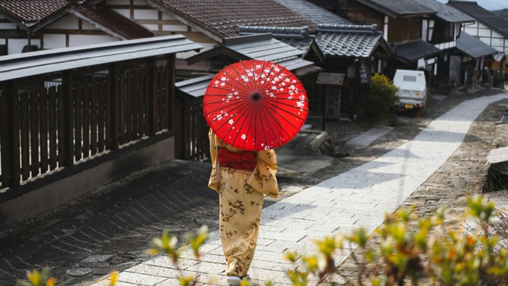 古き良き日本の街並みをゆったりと歩く女性。彼女は華やかな着物を着て、白い花柄の赤い和傘を差しています。背景には伝統的な家屋が並び、古い街の趣を感じさせます。 そして、iMessageとは？SMSとの違いをわかりやすく解説【日本版】