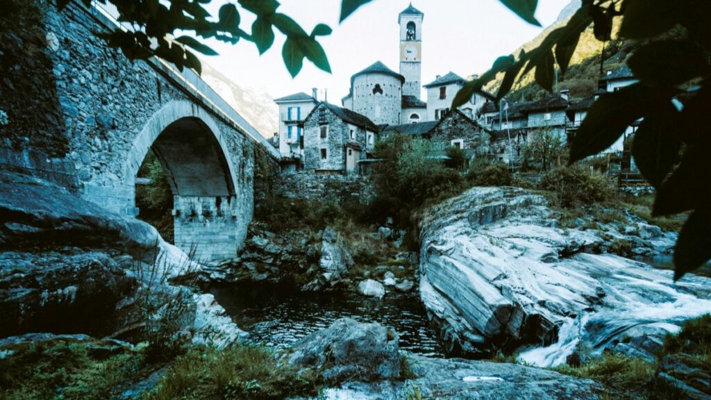 Un pueblo pintoresco con una estructura de piedra y un campanario en primer plano. Un arco de piedra cruza un arroyo, rodeado de enormes rocas y vegetación. La combinación de la naturaleza y la arquitectura antigua crea una escena encantadora. Además, ¿Cómo ganar dinero en Instagram desde España en 2025