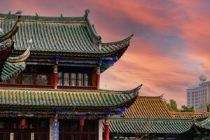 Este es un impresionante edificio de estilo arquitectónico chino tradicional, con techos curvados y techas de tejas verdes y amarillas. En el fondo, el cielo se ve repleto de nubes coloridas al atardecer. Además, una pregunta interesante para reflexionar: ¿Cómo pueden los influencers de facebook colaborar con marcas chinas?