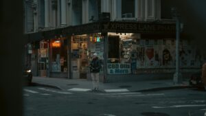 A street - corner view of an "Xpress Lane Pharmacy" at dusk, with illuminated signage and various advertisements in the windows, including "CBD Oils & More" and "PRESCRIPTIONS". A person stands on the sidewalk, adding a sense of everyday urban life. This image relates to the 2025 Charging Standards for TikTok Influencer Promotions in the U.S., capturing a typical city - side scene relevant to influencer marketing contexts.