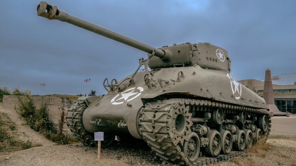 An old military tank with a weathered exterior and prominent white star insignias is displayed outdoors. The setting includes a gravel ground, some sparse vegetation, and a building with a pointed structure in the background. Flags can be seen flying in the distance under a cloudy sky. This image relates to the 2025 Charging Standards for Zalo Influencer Promotions in the U.S., highlighting a historical and visually striking subject relevant to influencer marketing contexts.