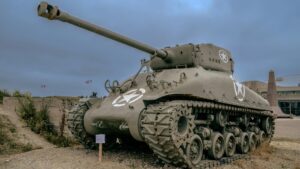 An old military tank with a weathered exterior and prominent white star insignias is displayed outdoors. The setting includes a gravel ground, some sparse vegetation, and a building with a pointed structure in the background. Flags can be seen flying in the distance under a cloudy sky. This image relates to the 2025 Charging Standards for Zalo Influencer Promotions in the U.S., highlighting a historical and visually striking subject relevant to influencer marketing contexts.