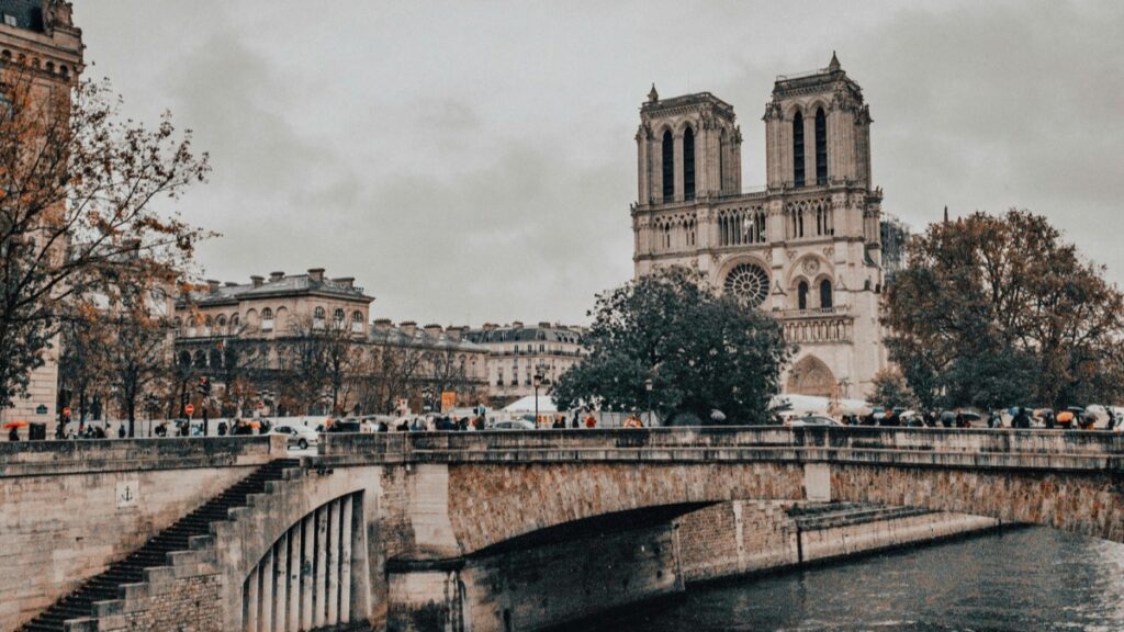 Une vue emblématique de Notre - Dame de Paris, majestueuse face au ciel gris. Le célèbre pont traverse la rivière, avec des passants qui s'y promènent, certains tenant des parapluies. Cette image incarne l'élégance parisienne et est en harmonie avec les 2025 Standards de Tarification pour les Collaborations avec des Influenceurs sur LINE en France.