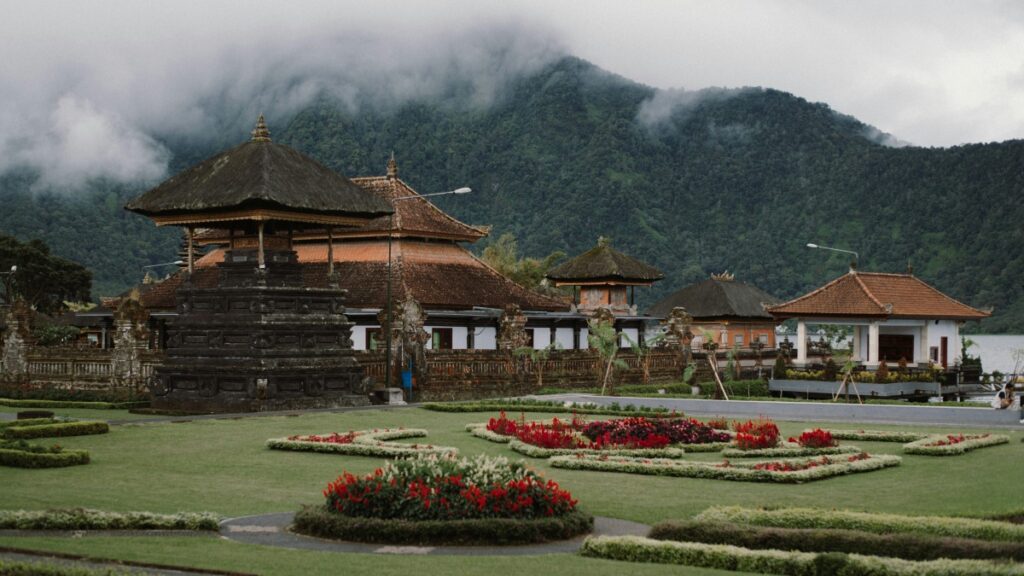 Gambar ini menunjukkan kompleks bangunan tradisional Bali dengan atap yang indah dan ukiran yang rumit. Di depan, ada taman yang teratur dengan bunga - bunga berwarna yang mewarnai lahan hijau. Di latar belakang, gunung yang ditutupi awan menambah pesona alam. Selain itu, jika Anda ingin mengetahui Cara Menggunakan WeChat untuk Pemasaran Bisnis di Indonesia, ada panduan yang dapat membantu Anda memanfaatkan platform tersebut untuk tujuan bisnis.